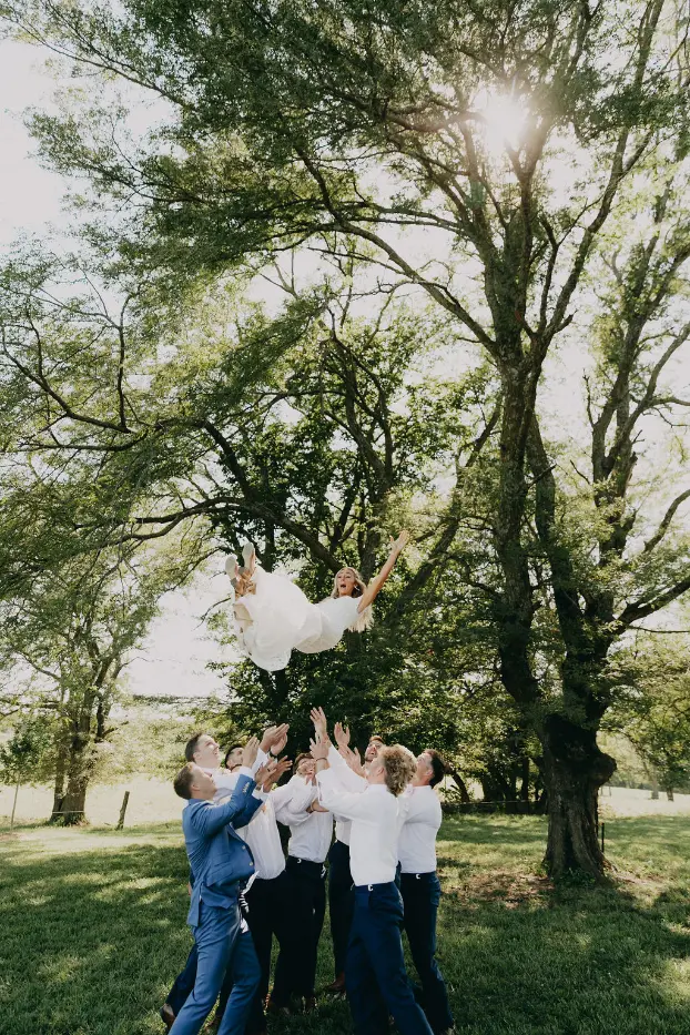 24 sarah and tyler at our kansas city wedding barn Bride being joyfully tossed in the air by groomsmen under lush trees at The Pearl wedding venue, showcasing a vibrant outdoor celebration.
