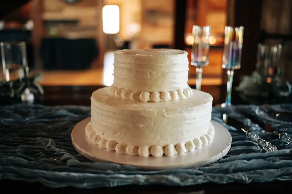 30 the bakers say i do at the pearl Two-tiered white wedding cake with decorative frosting and pearl accents, set on a textured blue table runner, surrounded by elegant glassware and a warm, inviting venue ambiance.