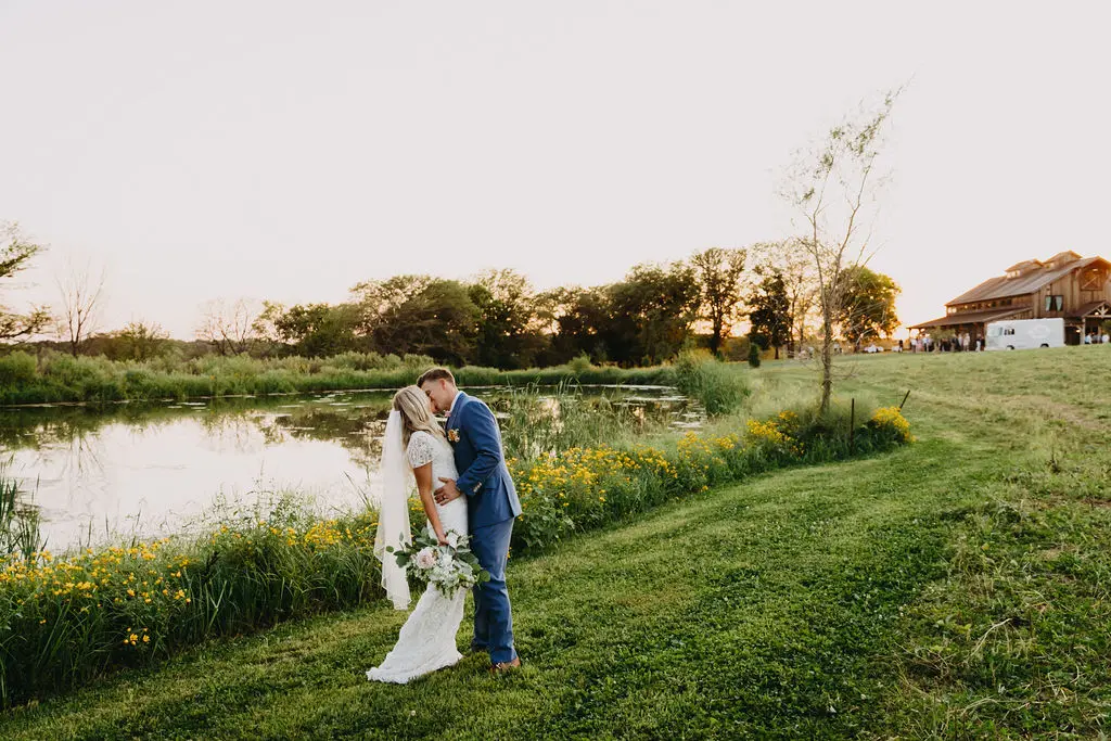 64 sarah and tyler at our kansas city wedding barn Bride and groom sharing a kiss by a serene pond at sunset, surrounded by lush greenery and wildflowers, with The Pearl at Crawford Farm venue in the background, highlighting a romantic and intimate wedding setting.