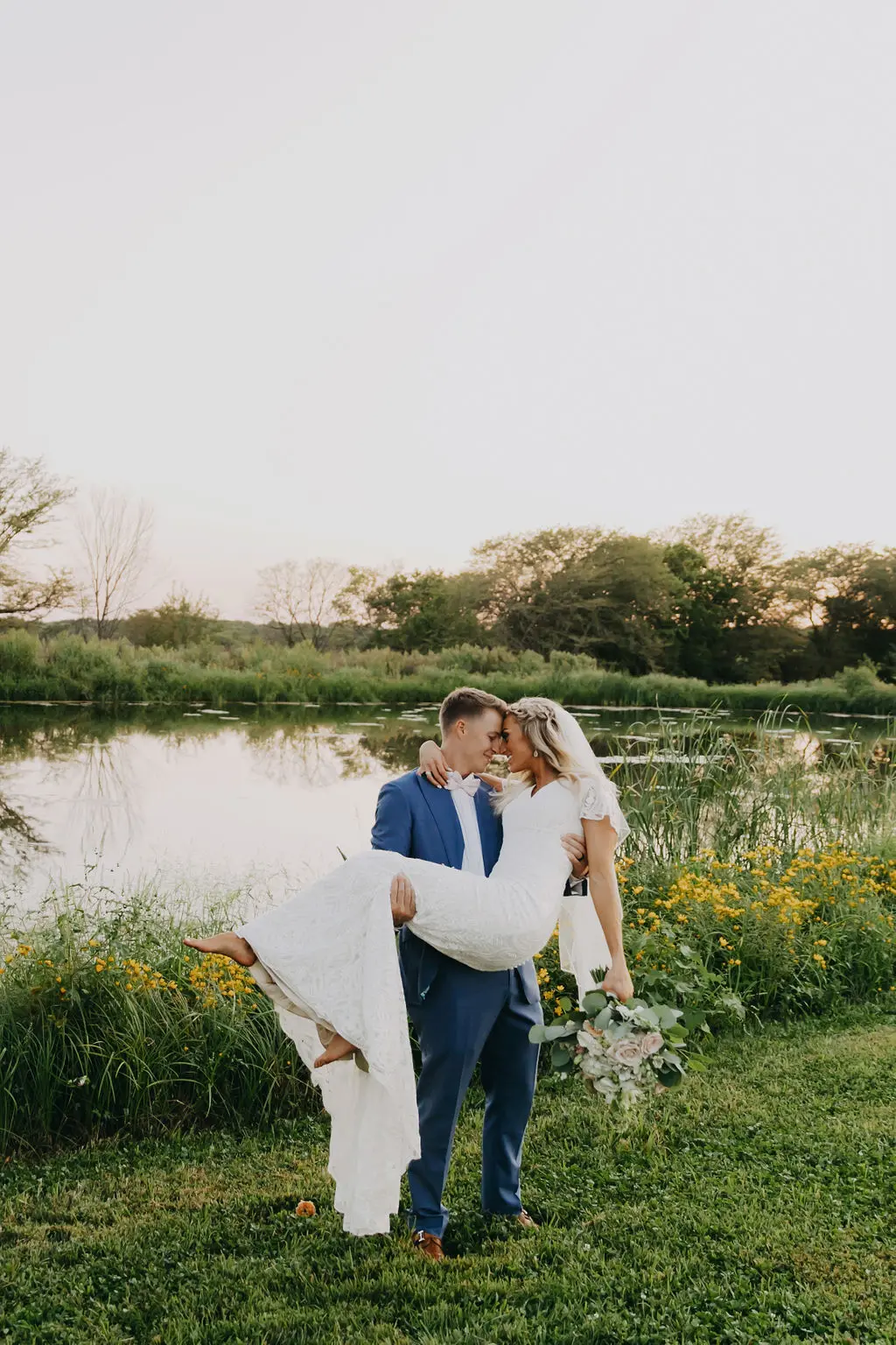 Bride in a lace wedding dress and groom in a blue suit sharing a romantic kiss near a pond, surrounded by greenery and yellow flowers, capturing the essence of a beautiful wedding at The Pearl at Crawford Farm in Manhattan, KS.
