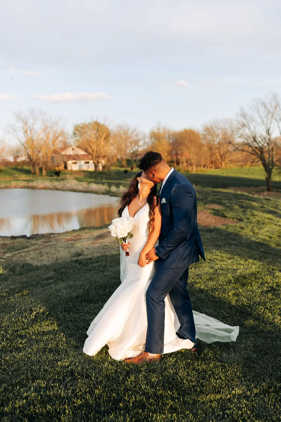 771 the bakers say i do at the pearl Bride and groom sharing a kiss by a serene pond at The Pearl at Crawford Farm, surrounded by rustic charm and natural beauty, showcasing a romantic wedding setting.