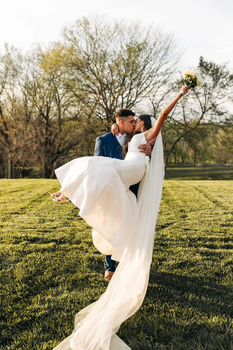 781 the bakers say i do at the pearl Bride and groom embracing in a romantic kiss, with the bride holding a bouquet and her wedding veil flowing, set against a lush green landscape at The Pearl wedding venue in Missouri.