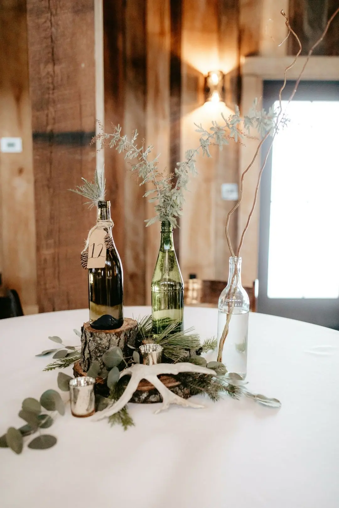 Winter wedding table centerpiece featuring wine bottles, pine branches, and eucalyptus on a rustic wooden slice, enhancing cozy lodge vibes.