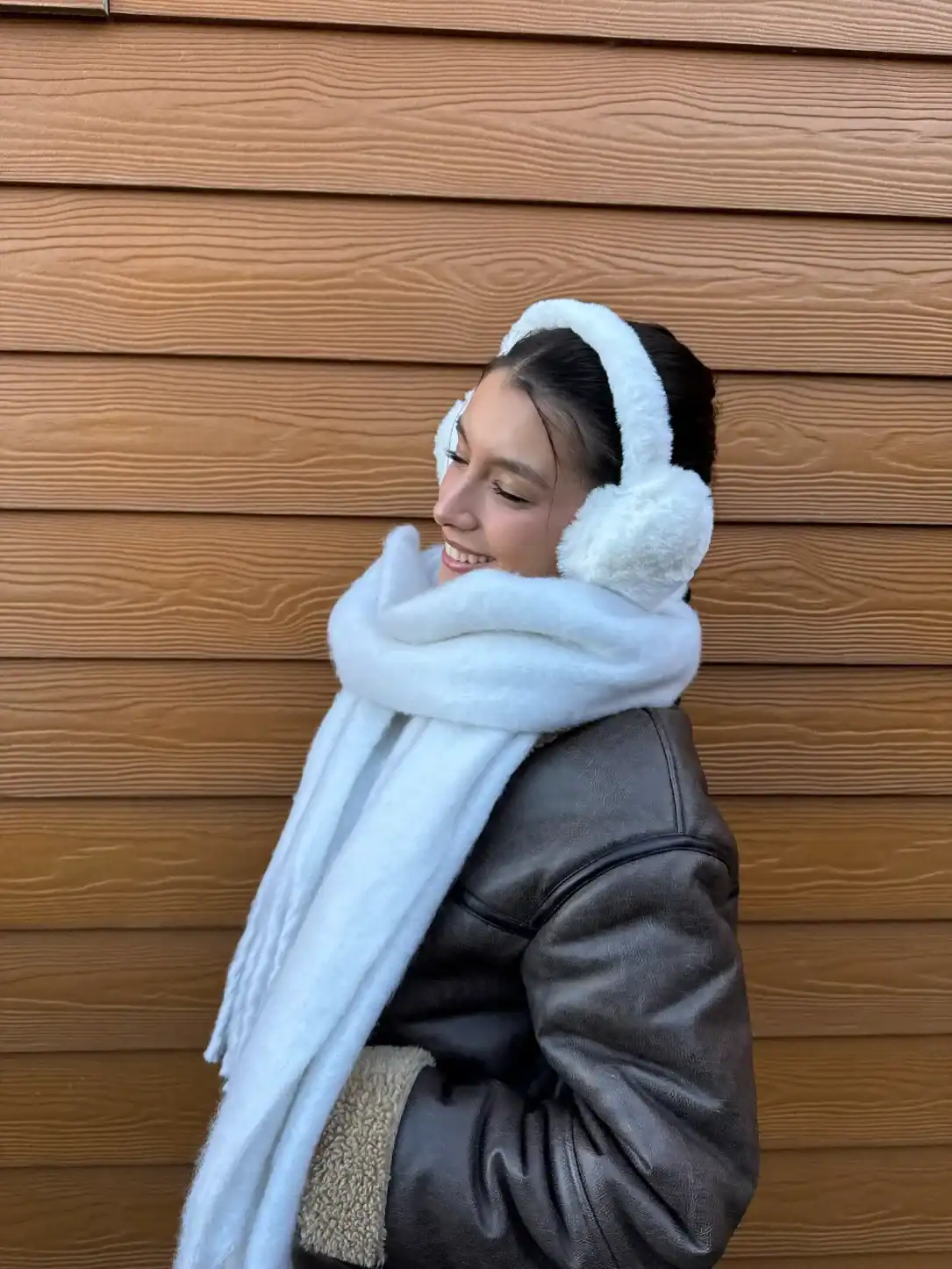 Young woman smiling in winter attire, wearing fluffy earmuffs and a cozy scarf, against a wooden backdrop, embodying the warmth and style of winter weddings.