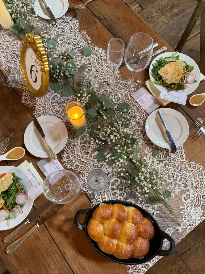 Elegant wedding reception table setting with lace runner, candles, and greenery, featuring a golden table number, glassware, and plates of salad and warm bread rolls, embodying winter wedding ambiance and comfort food themes.