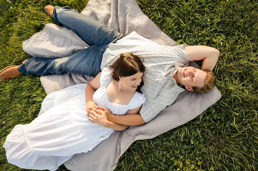 Couple lying on a blanket in a grassy field, smiling and enjoying a relaxed moment together, representing the romantic atmosphere ideal for engagement photos at The Pearl at Crawford Farm wedding venue.