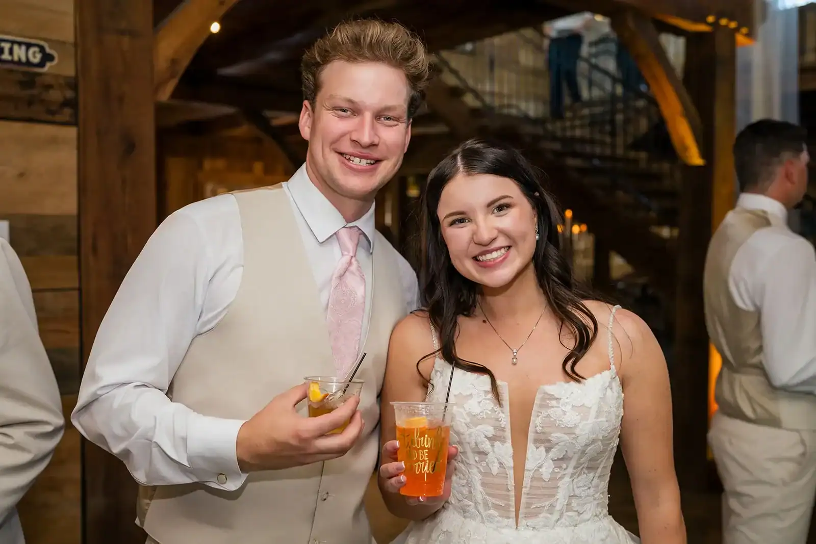 Happy couple celebrating at The Pearl wedding venue, dressed in formal attire, holding drinks, with rustic barn decor in the background.
