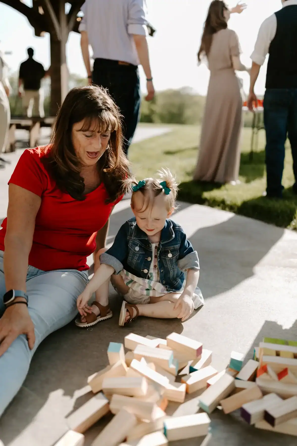 Woman and child playing with colorful wooden blocks on a paved surface at a wedding venue, with guests mingling in the background.