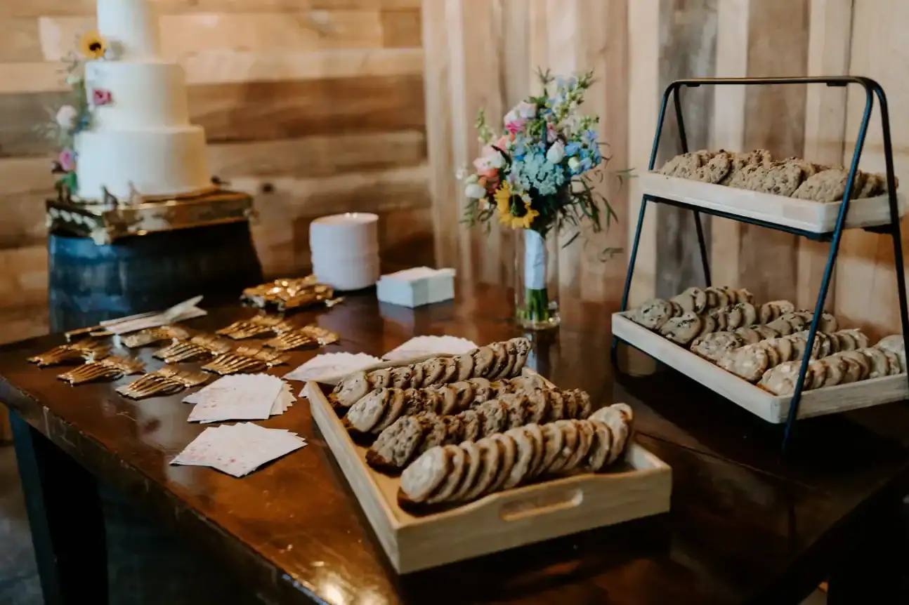 Dessert table featuring assorted cookies and a three-tier wedding cake, with floral arrangement and decorative utensils, set in a rustic venue.