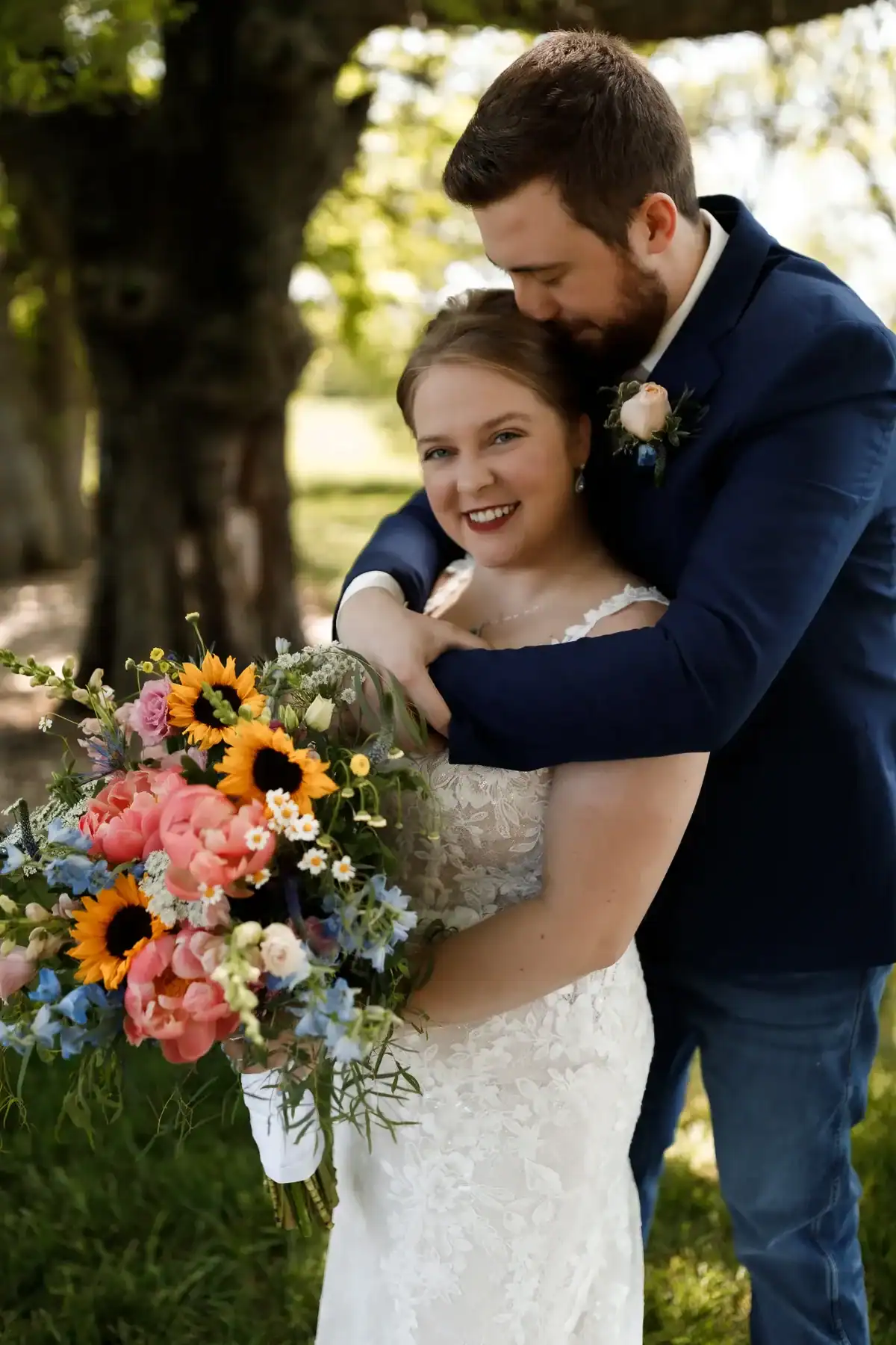 Bride and groom embracing in outdoor setting, bride holding vibrant bouquet of sunflowers, peonies, and wildflowers, showcasing love and celebration for weddings at The Pearl at Crawford Farm in Topeka, KS.