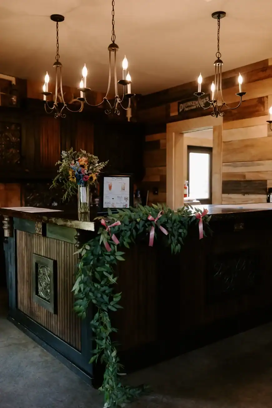 Rustic bar area with elegant chandeliers, floral arrangement, and greenery accents, showcasing a welcoming atmosphere for events at The Pearl at Crawford Farm.