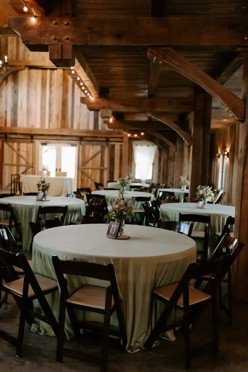 Rustic barn interior featuring round tables with green tablecloths, floral centerpieces, and wooden chairs, ideal for weddings and events at The Pearl at Crawford Farm in Topeka, KS.